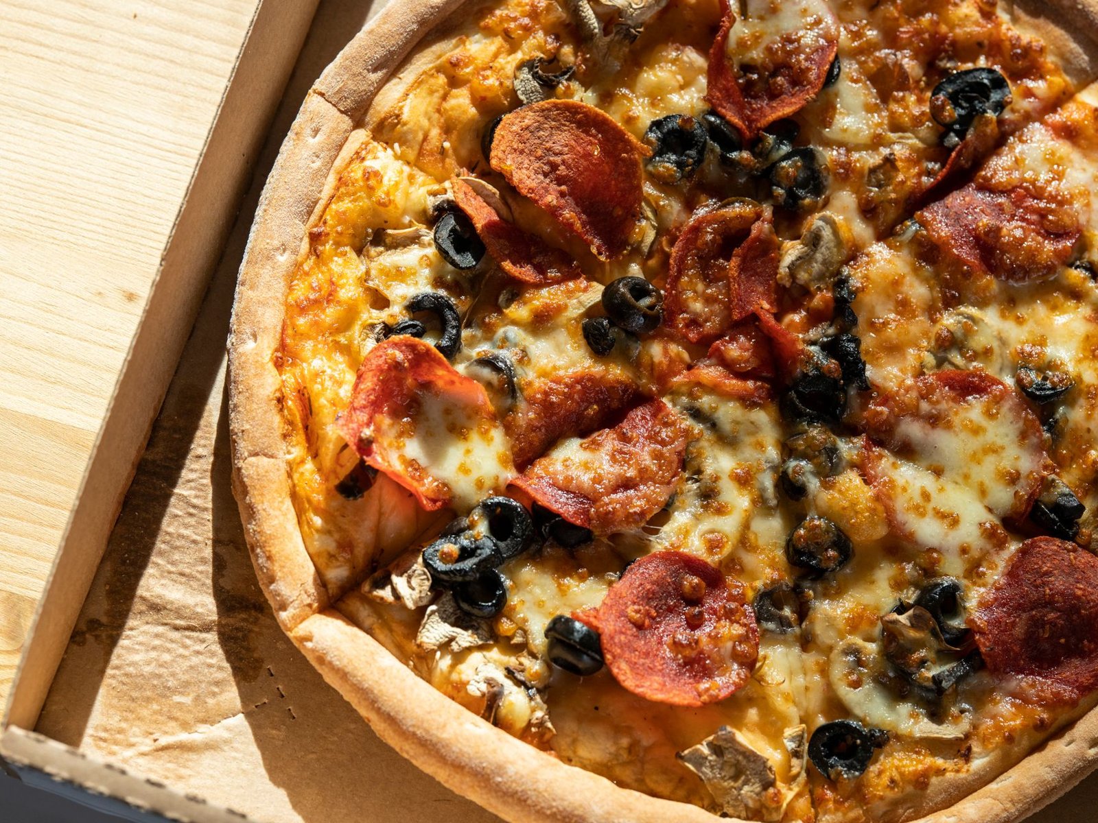 Pizza tonda con salame piccante, olive nere, funghi e mozzarella fusa in un cartone da asporto su tavolo di legno